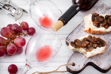 2 bardak, kırmızı şarap, tirbuşon, üzüm, beyaz ahşap arka planda peynirli ve mantarlı aperatifler.