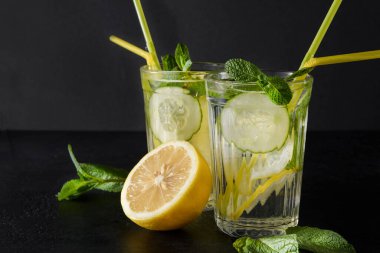 Sarı limonlu 2 bardak limonata, siyah arka planda taze salatalık, yeşil nane ve tüpler, kopyalama alanı.,