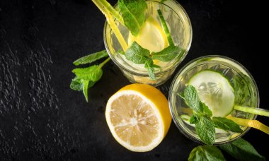 Sarı limonlu 2 bardak limonata, taze salatalık dilimleri, siyah arka planda yeşil nane ve tüpler, fotokopi alanı, üst manzara