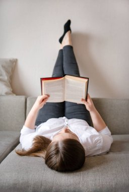 Bir beyaz gömlekli beyaz bir kadın kanepede uzanmış kitap okuyor.