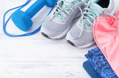 Gri spor ayakkabılar, 1 mavi dambıl, atlama ipi, pembe bluz, beyaz ahşap arka plan manzaralı taytlar.