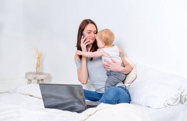 Evde çalışmak, beyaz bir anne yatakta bebekle oturup telefonla konuşmak ve dizüstü bilgisayarla çalışmak., 