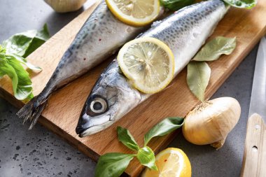 Kesme tahtasında baharatlı ve limon dilimli 2 çiğ uskumru balığı, fesleğen, sarımsak, soğan, gri arka planda bıçak.