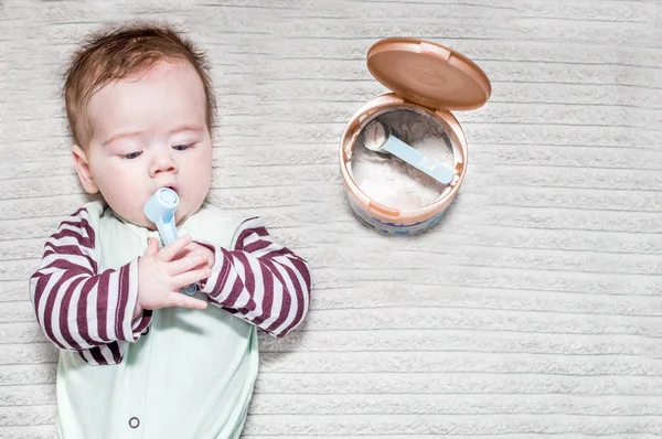 Bebé está acostado con una cuchara en la mano cerca de un frasco de fórmula infantil — Foto de Stock