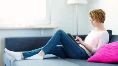 güzel kadın gri kanepede yatıyor ve tablet üzerinde çalışır