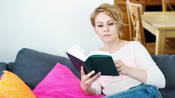 美丽的女人坐在灰色的沙发上看书在日光下 — 图库视频影像