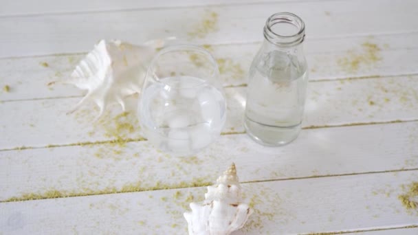 Glas mit Wasser und weißen Muscheln auf Holztisch mit Sand — Stockvideo