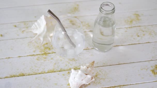 Verre avec eau avec paille de titane et coquillages blancs sur table en bois avec sable — Video