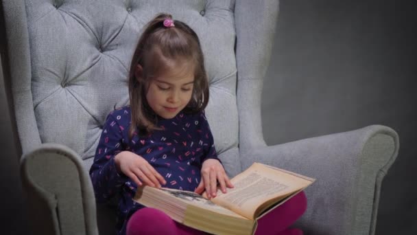 Adorável menina olha através de um livro na cadeira — Vídeo de Stock