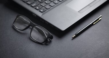 Siyah arkaplanda dizüstü bilgisayar, kalem ve gözlük var..