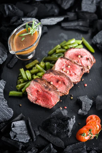 Medalhão de bife com legumes e molho em uma placa preta com carvão — Fotografia de Stock