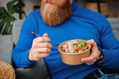 Bir adam evinde, kanepede yemek yiyor. Mavi kazaklı adam. Ev yemeği..