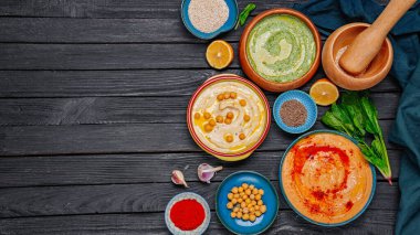 Renkli humus kaseleri, siyah ahşap arka planda sağlıklı vegan sosları. Ispanaklı humus, kırmızı biber ve zira. Geleneksel Ortadoğu humusu. Sağlıklı beslenme konsepti. Ispanaklı humus, kırmızı biber ve zira.