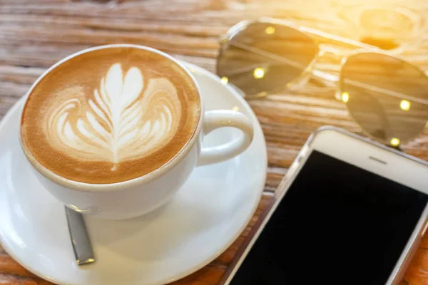 Taza de café arte latte con cuchara y plato en la corteza marrón hermoso fondo textura con luz cálida decorada con gafas de sol — Foto de Stock