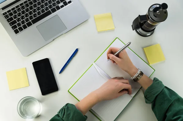 Escrevendo no bloco de notas sentado na mesa com smartphone e computador portátil — Fotografia de Stock