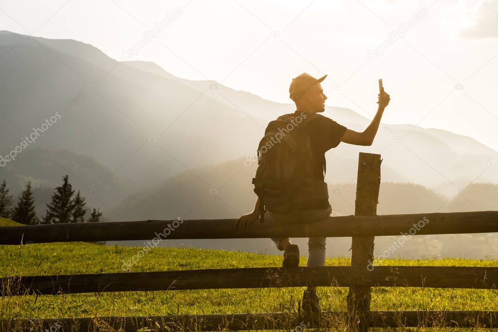 Young male hiker sits on fence in ukrainian carpathian mountains and communicates by mobile phone or takes self portrait