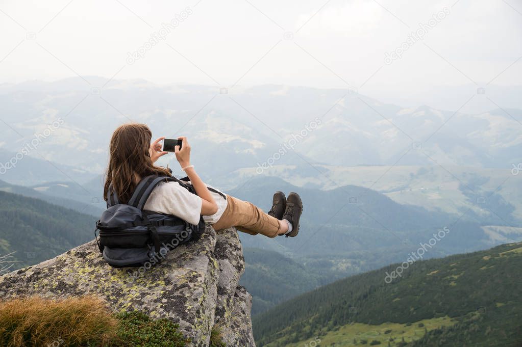 Female tourist takes photo of legs with cell phone from top of the mountain cliff