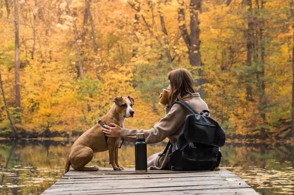 Caminhante turístico menina senta-se na margem do rio com seu filhote de cachorro de estimação e goza de bela vista indiana de verão com xícara de café de garrafa térmica — Fotografia de Stock