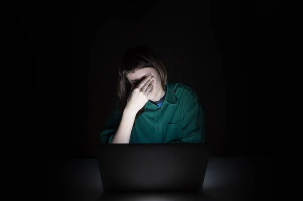 Tired and exhausted young woman rubs eyes at laptop pc late in the evening