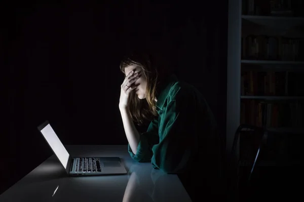 Усталая и измученная молодая женщина закрывает глаза рукой за ноутбуком поздно вечером — стоковое фото