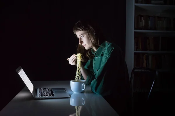 Giovane femmina al computer portatile mangia spaghetti istantanei ramen con bacchette a tarda sera — Foto Stock