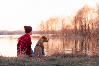 Genç adam ve evcil köpek bahar veya sonbahar günbatımı göl kenarında oturup