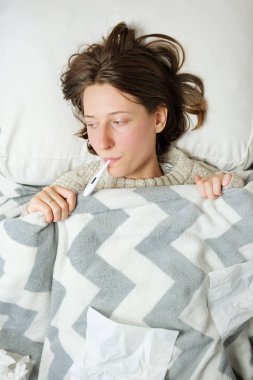 Genç bir kadın. Yatakta bir battaniyenin altında ağzında termometreyle yatıyor.. 