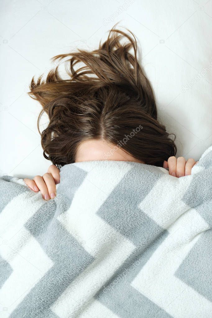 A young woman lies in bed under a blanket and have a hard time waking up in the morning.