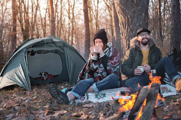 Two persons enjoying hot drinks by the bonfire at a camping site.