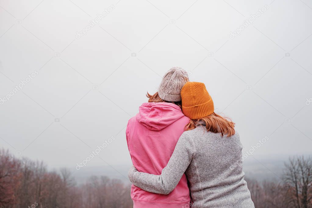 Two young woman in casual clothes hug each other on autumn sky background. 