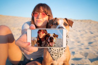 Human taking a selfie with dog. Best friends concept: young female makes self portrait with her puppy outdoors on a beach clipart