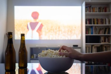 Ev sineması eğlencesi: bir odadaki video projektöründen film izlemek. Loş oturma odası, duvarında sinema resmi olan, insan eli patlamış mısır tutan.