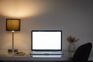 Geceleri rahat ofis masası bir lamba ve beyaz dizüstü bilgisayarla aydınlatılıyor. Tele-iş ya da serbest faaliyet için temiz ve modern bir iş yeri, gösterişsiz bir ortam.