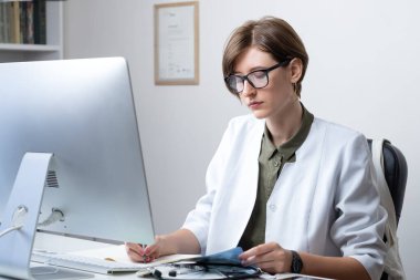 Modern tıp muayenehanesinde çalışan kadın pratisyen. Genç tıp doktoru iş yerinde bilgisayarın önünde notlar alıyor.