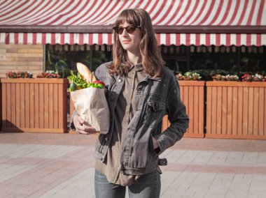 Yerel sebze ve marketten ya da marketten alınmış bakkaliyeden alınmış günlük kıyafetleri olan çekici genç bir kadın. Geri dönüşümlü sağlıklı vejetaryen yiyecekler kadın ellerinde..