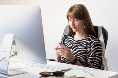 Young female worker at desk in modern minimalistic office. Portrait of business woman using mobile phone in front of desktop computer
