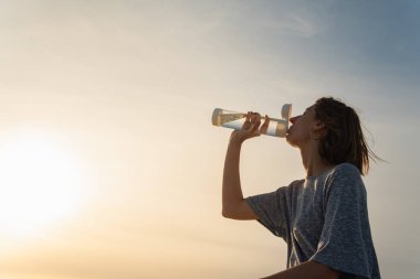 Genç bir kadın, dışarıda parlak güneş ışığıyla şişeden su içiyor.