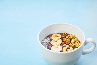 Bir kase fındıklı ve muzlu meyve püresi. Mısır gevreği, kaju ve fındıklı Acai kasesi mavi parlak arka planda.