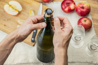 Erkek ellerinin prim şişesini açtığının en üst görüntüsü. Yukarıdan çekilen güzel buzlu soğuk elma şarabı şişesi arka planda yetişmiş olgun elmalar.
