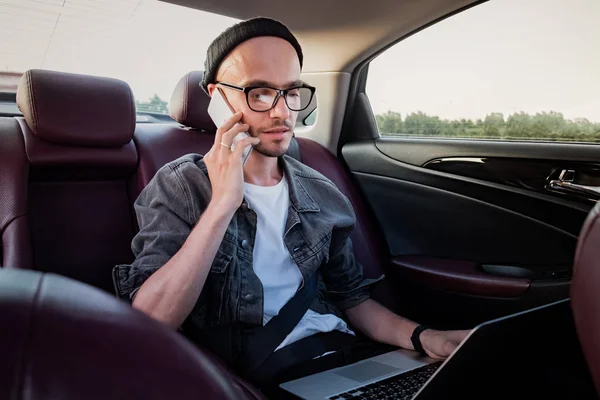 Man with laptop making phone call on a backseat of a car on trip to work. Male person using modern technology while commuting by a private or rental car