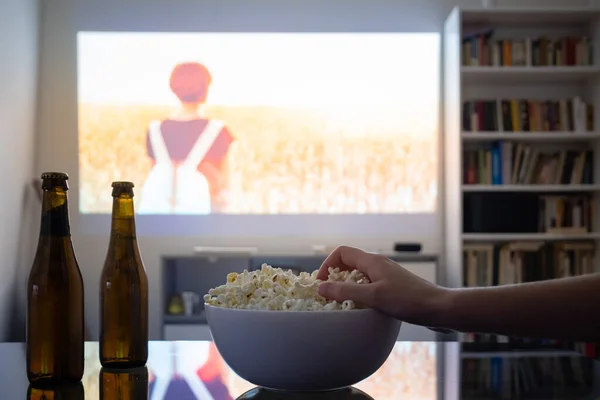 Ev sineması eğlencesi: bir odadaki video projektöründen film izlemek. Loş oturma odası, duvarında sinema resmi olan, insan eli patlamış mısır tutan.