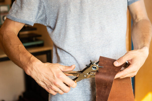 Making holes with puncher in leather workshop. Male hands with eyelet puncher working with piece of material