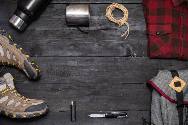 Preparing for a hiking trip: backpacking things and clothes flat lay. Top view of trekking boots, tourist rucksack and camping acessories on black background