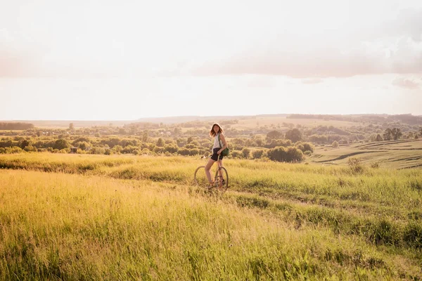 Girl with a bicycle posing on the hill in beautiful rural landscape at sunset. Young pretty female person with retro bike standing in a meadow on bright sunny afternoon in summer