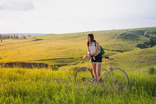 Girl with bicycle taking a rest in beautiful rural landscape. Young pretty female person with retro bike standing in a meadow on bright sunny afternoon in summer