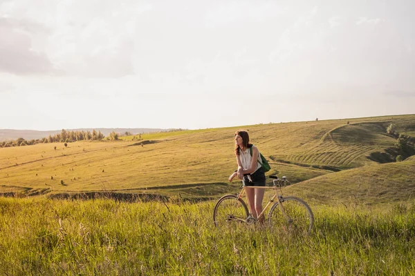 Girl with bicycle taking a rest in beautiful rural landscape. Young pretty female person with retro bike standing in a meadow on bright sunny afternoon in summer