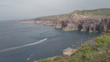 Körfeze ulaşan beyaz bir motorlu tekne, kayalıklar ve vahşi doğa arasındaki nefes kesen bir manzarada koyu mavi deniz suyuyla kontrastlı beyaz köpük izi.