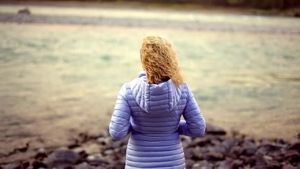 Женщина в синей куртке и солнцезащитных очках простирает руки возле горной реки, каменного пляжа — стоковое видео