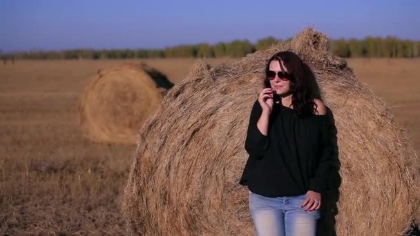 Flicka i en svart jacka och solglasögon som står nära höstack — Stockvideo