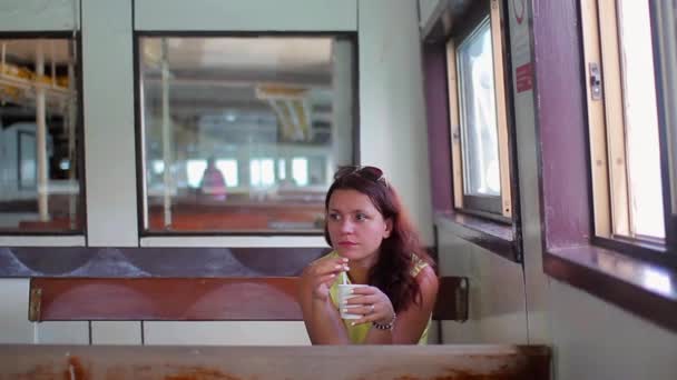Uma menina em um vestido amarelo sentado em um banco em ferry de carga, bebe café de uma xícara de papel e olhando pela janela — Vídeo de Stock